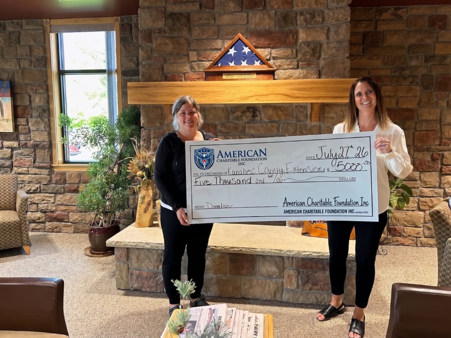 Two women presenting a large ACF grant check for Kanabec County Extension in a stone-walled lobby