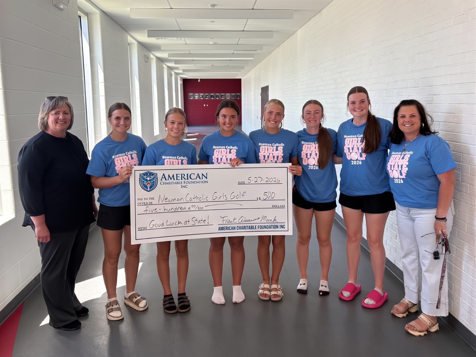 Two adults and seven students in matching blue shirts presenting a large ACF grant check in a school hallway