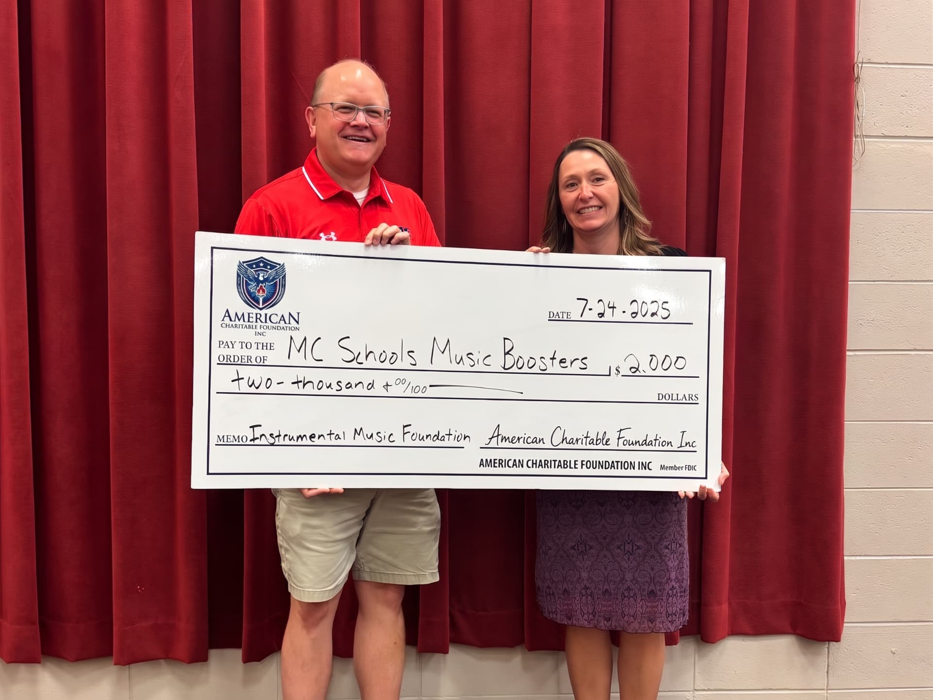 Two people presenting a large ACF grant check for MC Schools Music Boosters in front of red stage curtains